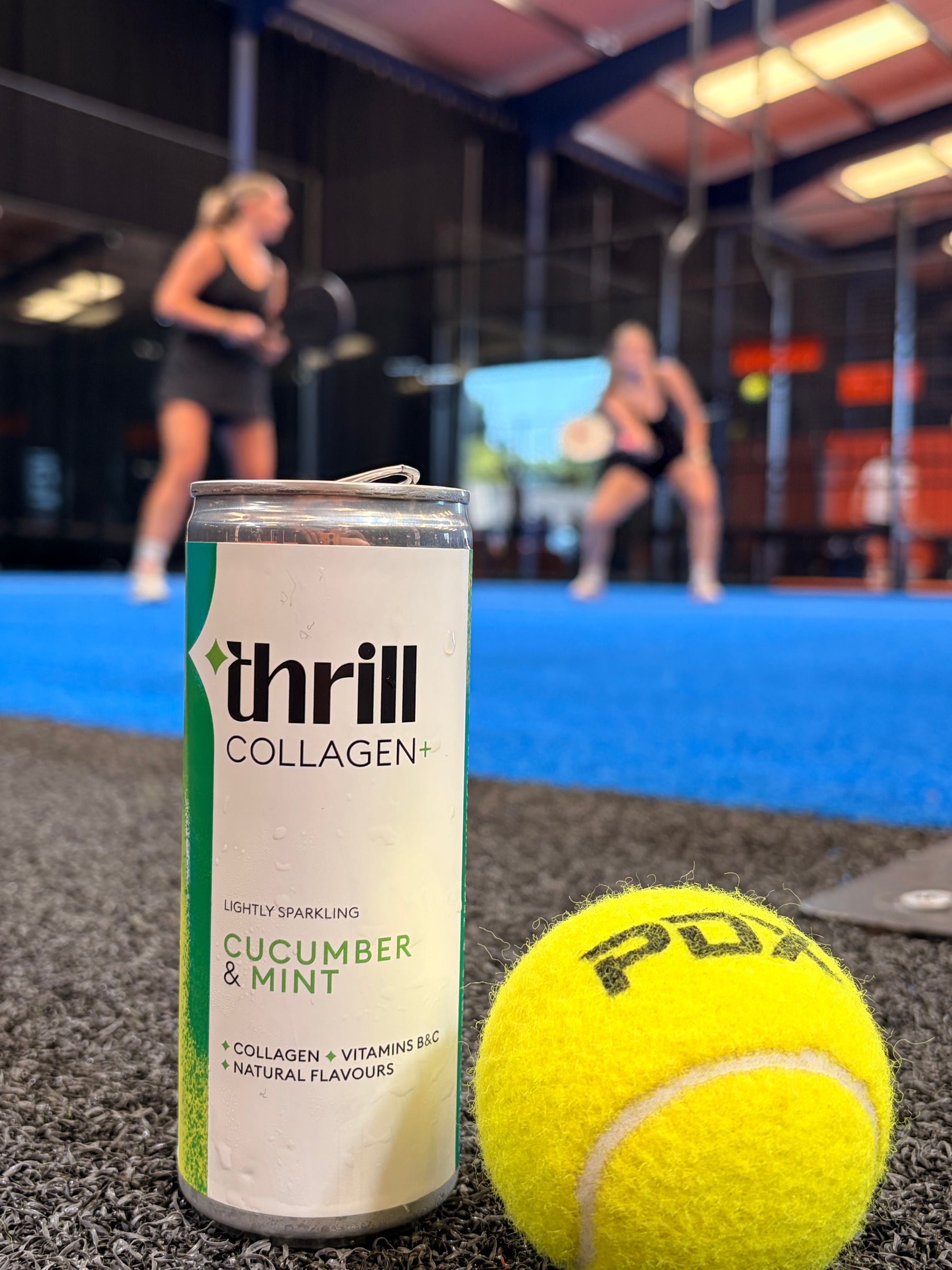 A can of Thrill Collagen cucumber and mint drink in the foreground, next to a yellow tennis ball, with a blurred indoor sports court and people in the background.